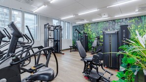 A gym with various exercise equipment, large windows, and a green leafy wall.