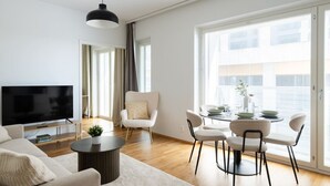 A modern living room with a flat-screen TV, a round coffee table, a sofa, a dining area with a table and chairs, and large windows with blinds.