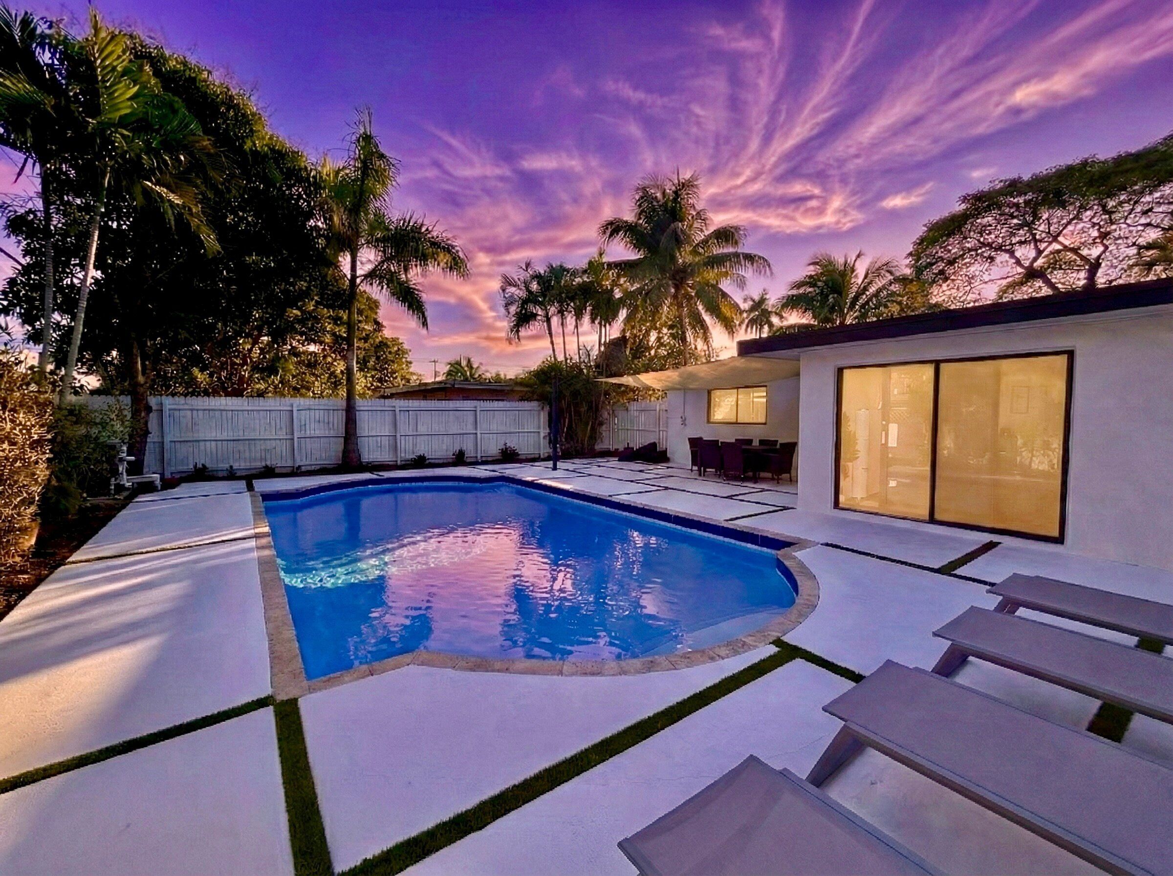 A swimming pool with a white deck, surrounded by palm trees and a white fence.