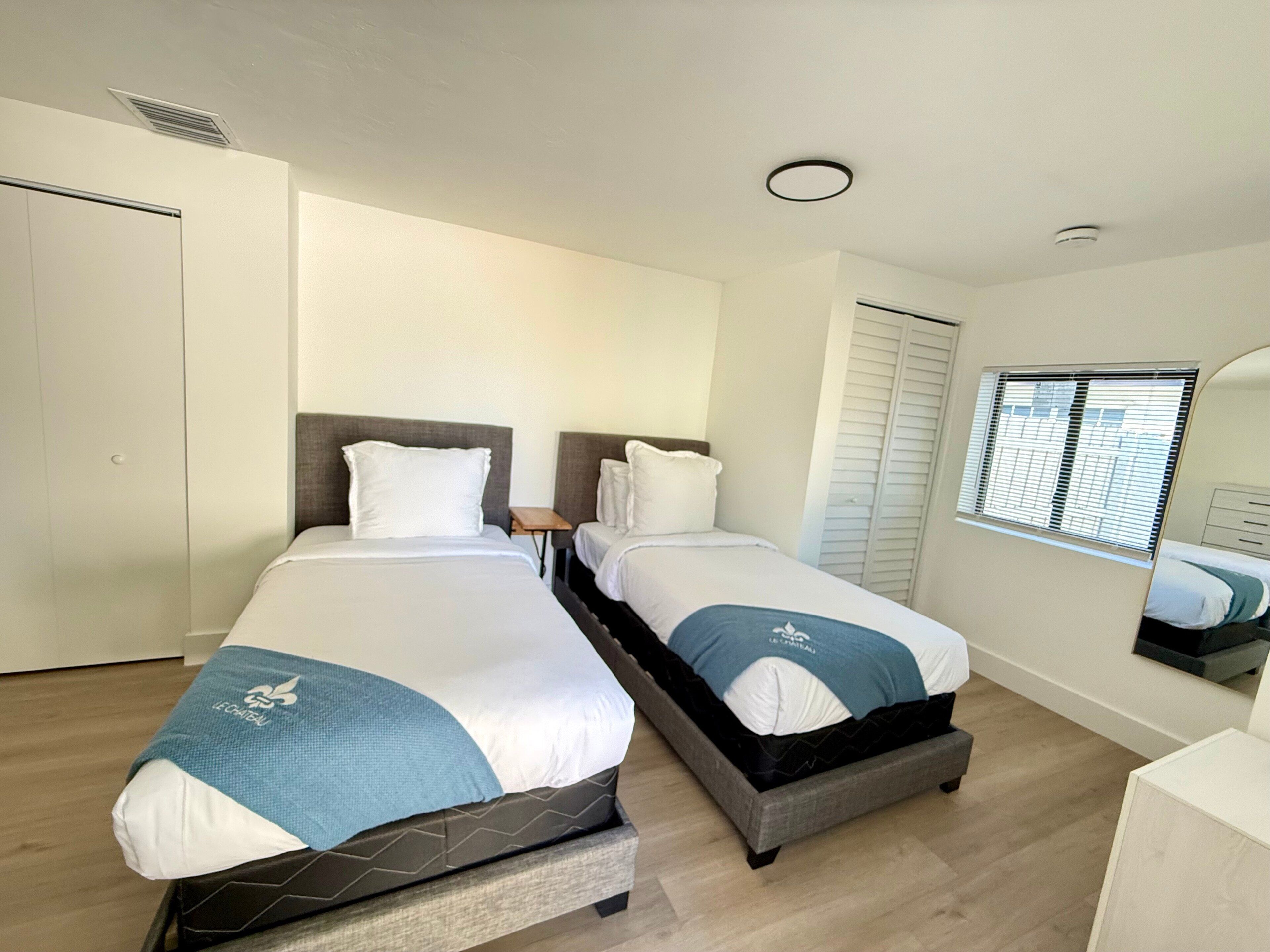 A hotel room with two beds, a wardrobe, a window with blinds, and a ceiling vent.