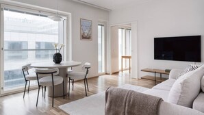 A modern living room with a white sofa, a round dining table with chairs, a flat-screen TV mounted on the wall, and large windows offering a view of the city. - By Tripla & Train Station | King Bed · 55 TV · Gym (Helsinki)