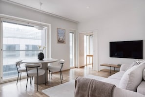 A modern living room with a white sofa, a round dining table with chairs, a flat-screen TV mounted on the wall, and large windows offering a view of the city. - By Tripla & Train Station | King Bed · 55 TV · Gym (Helsinki)