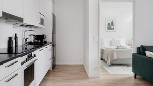 A modern kitchen with white cabinets, a black countertop, and stainless steel appliances. A bedroom with a bed, pillows, and a green sofa is visible through an open door. - Sauna, balcony, wifi, trainstation, Mall of Tripla (Helsinki)