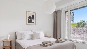 A bedroom with a bed, pillows, a bedside table, a lamp, a window with curtains, and a view of buildings. - Sauna, balcony, wifi, trainstation, Mall of Tripla (Helsinki)