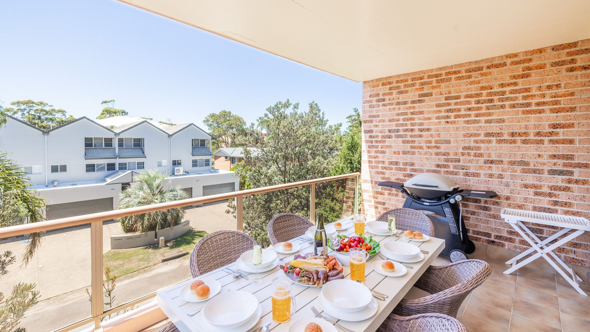 A balcony with a table set for a meal, a grill, and a view of buildings and trees.