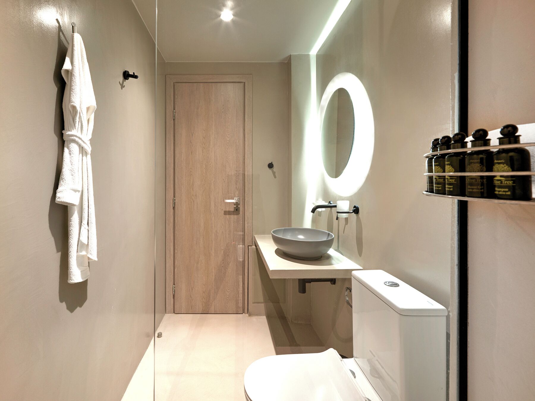 A modern bathroom with a wooden door, a round mirror, a white sink, and a shelf with toiletries.