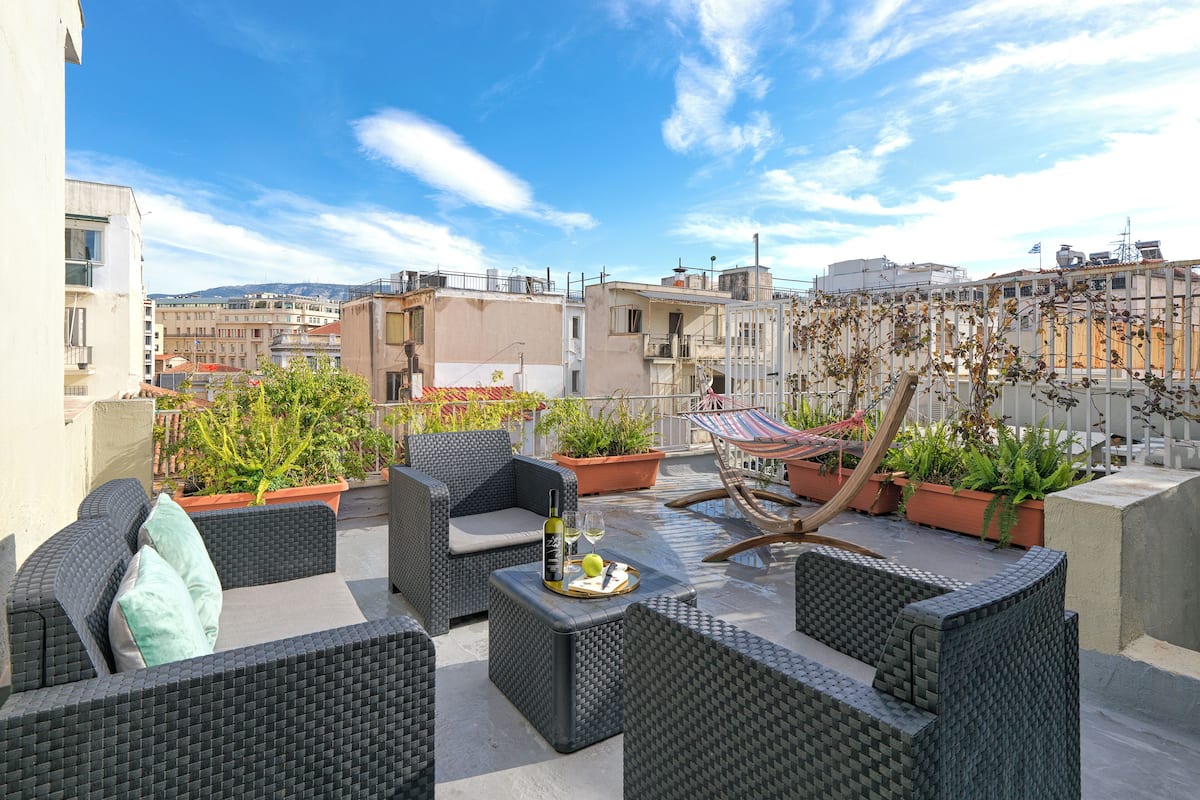 A rooftop terrace with wicker furniture, potted plants, and a glass table with a bottle and glasses.