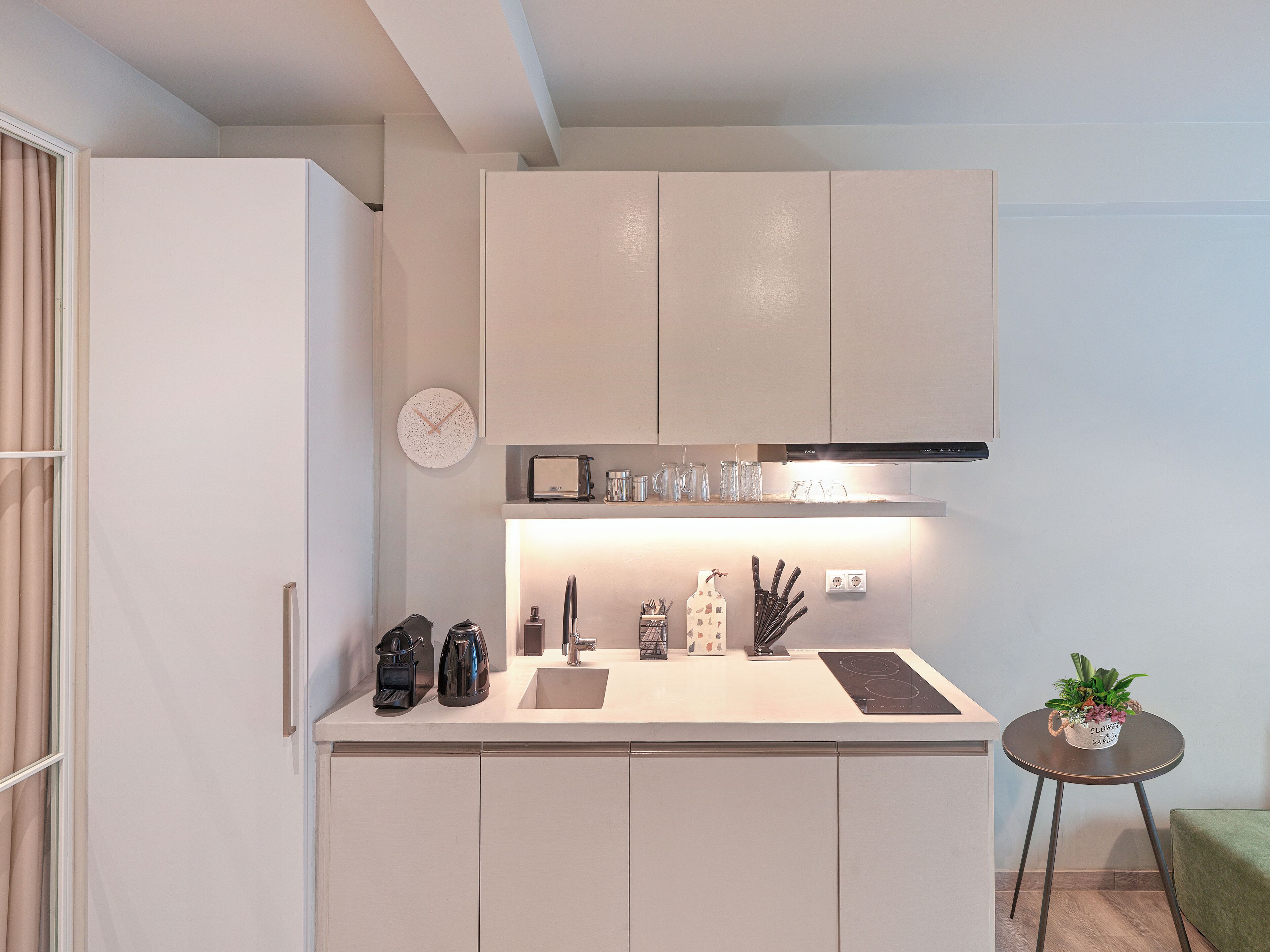 A compact kitchen with white cabinets, a built-in oven, a coffee maker, and a small table with a potted plant.