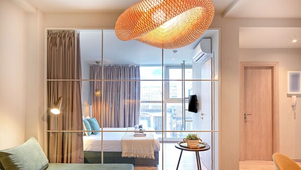 A modern hotel room with a large window, a green sofa, a small round table with a plant, and a unique orange pendant light. - Karitsi Suites (Athens)