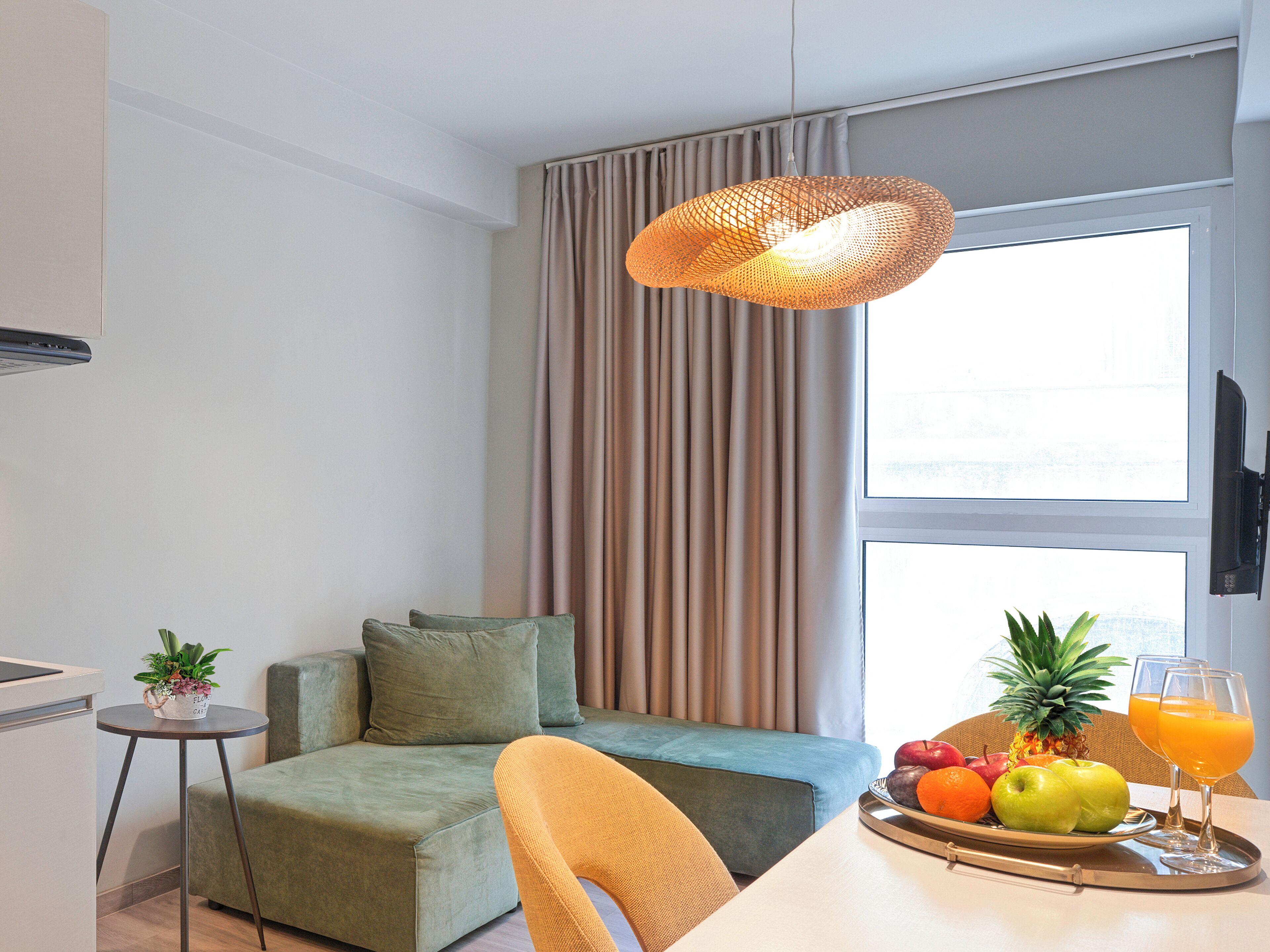 Une chambre d’hôtel moderne avec un canapé, une table à manger avec des fruits et du jus, un petit coin cuisine et une grande fenêtre avec des rideaux.