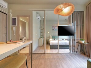 Una habitación de hotel moderna con una cama grande, un televisor, un escritorio con una silla, una mesita con una planta y un ventanal con cortinas.