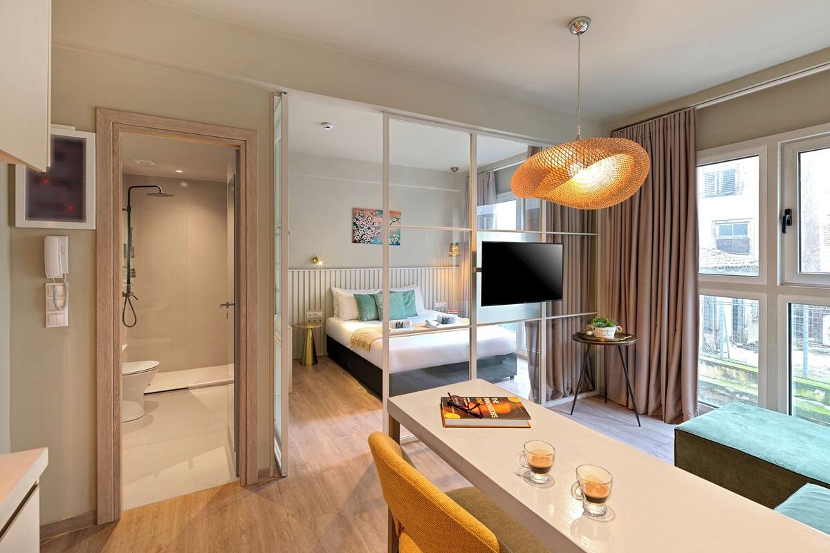 A modern hotel room with a glass partition separating the bedroom from the bathroom, a flat-screen TV, a dining area with a table and chairs, and a large window with curtains.