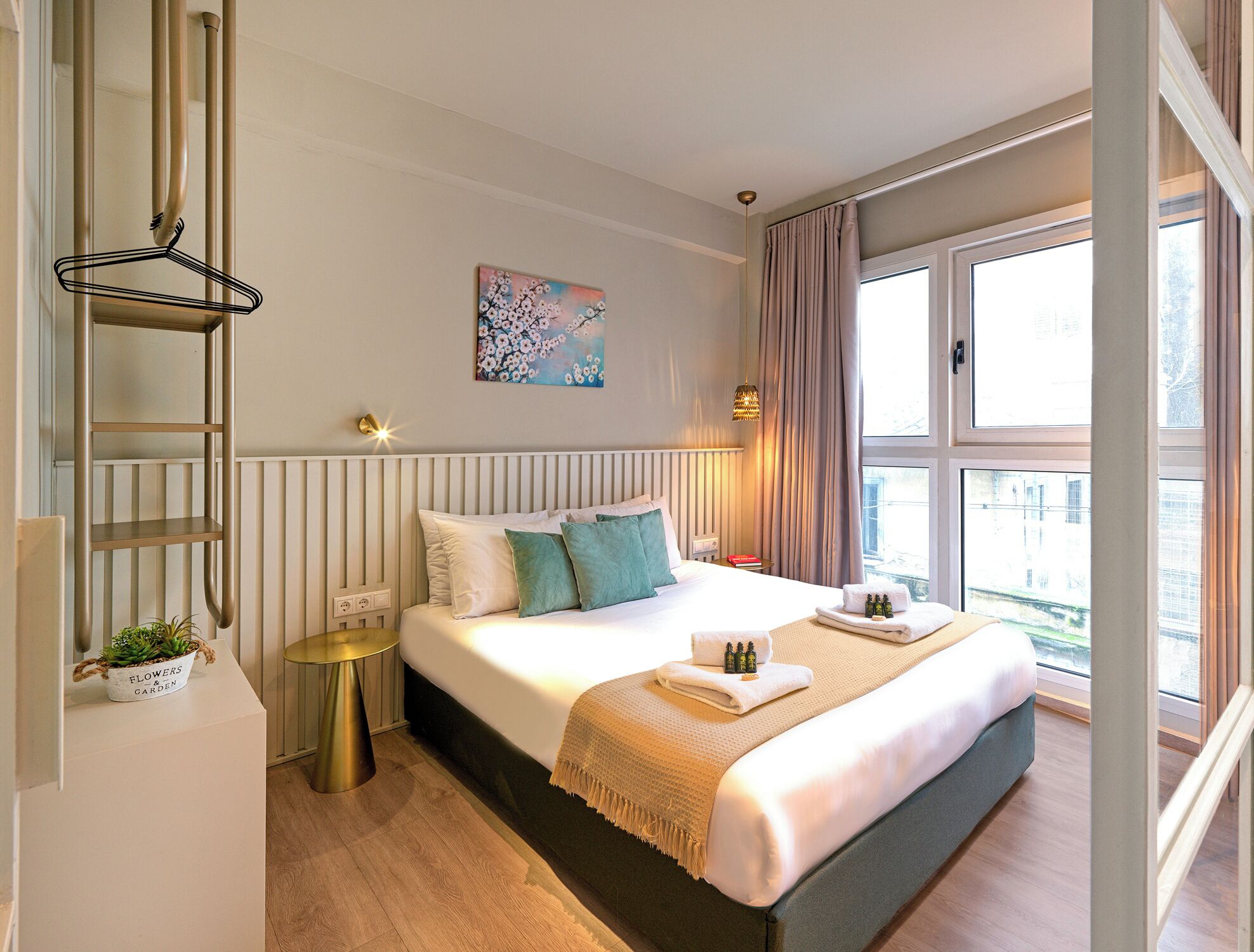 A modern bedroom with a large bed, a bedside table, a hanging light fixture, a window with curtains, and a wall-mounted shelf.