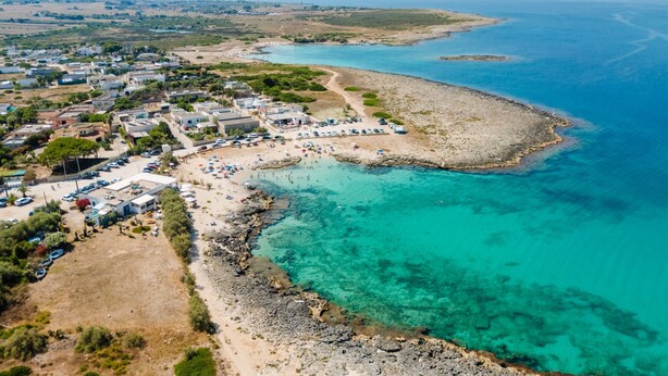 Une vue aérienne d’une ville côtière avec une plage, des eaux d’un bleu limpide et un mélange de bâtiments résidentiels et commerciaux.