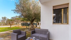 Un espace extérieur aménagé avec des meubles en osier, une table sur laquelle se trouve une assiette de biscuits et une tasse, et un grand arbre en arrière-plan.