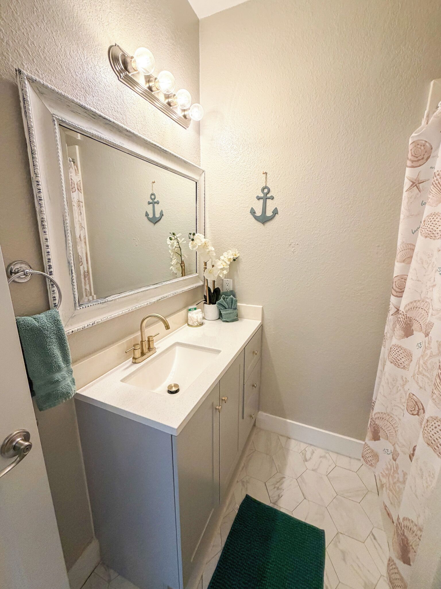 Une salle de bain avec un meuble-lavabo, un évier et un miroir. On y trouve des éléments de décoration en forme d’ancre au mur, ainsi qu’un rideau de douche orné d’un motif de coquillages.