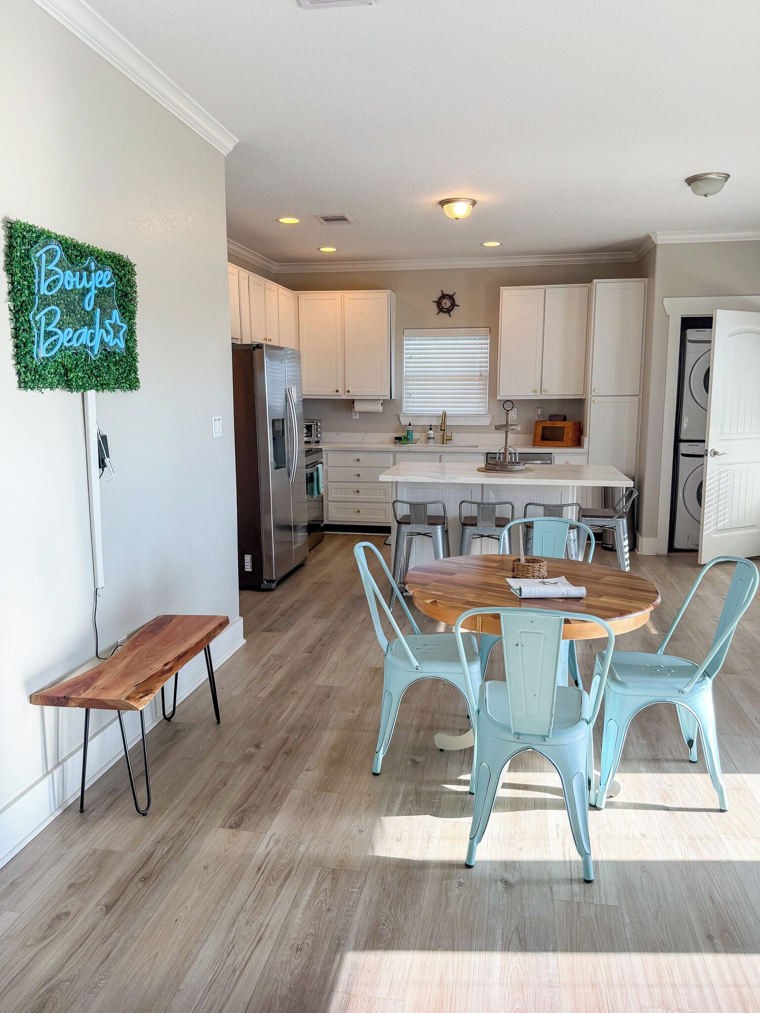 A modern kitchen with white cabinets, a wooden dining table with blue chairs, and a neon sign on the wall.