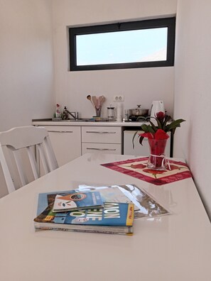 Ein Essbereich mit einem weißen Tisch, einer Zeitschrift, einer Vase mit roten Blumen und einem Fenster über der Arbeitsfläche.