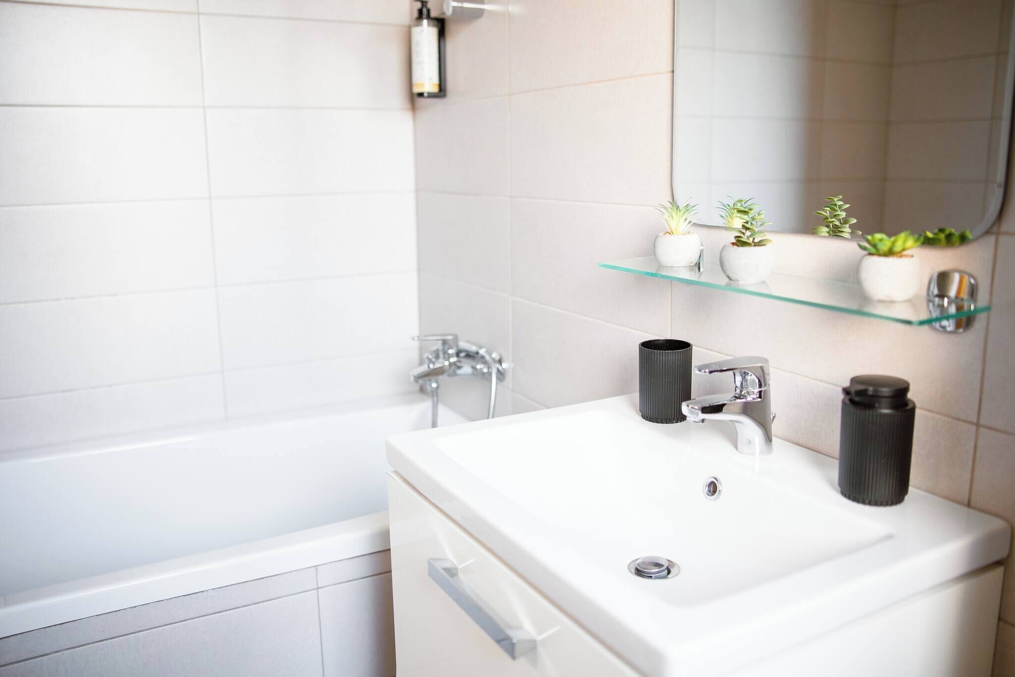 Ein modernes Badezimmer mit einem weißen Waschbecken, zwei kleinen Topfpflanzen auf einer Glasablage, einer schwarzen Vase und einer Dusche.