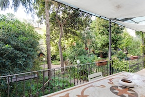 A covered outdoor area with a table and chairs, surrounded by greenery and trees.