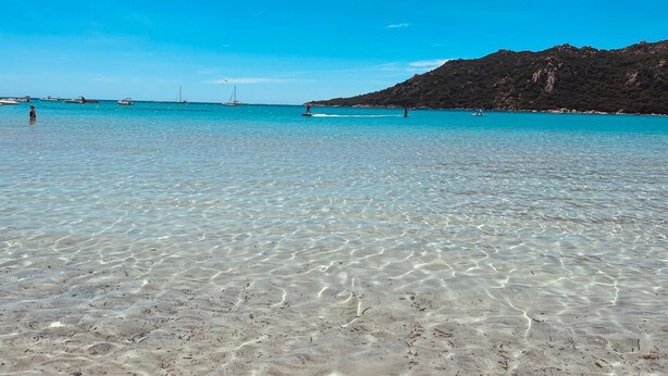 Una playa despejada con aguas poco profundas, barcos y montañas al fondo.