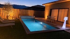 Una piscina con sillas de descanso y una mesa, rodeada por una valla de madera y un edificio con techo de tejas.