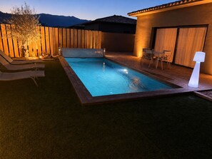 Ein Schwimmbecken mit Liegestühlen und einem Tisch, umgeben von einem Holzzaun und einem Gebäude mit einem Ziegeldach.