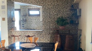 A dining area with a glass-top table, wooden chairs, a herringbone-patterned floor, a kitchen with a tiled backsplash, and a guitar leaning against the wall. - Holiday Home 'Dall'alba Al Tramonto' with Private Terrace, Wi-Fi and Air Conditioning (Alezio)