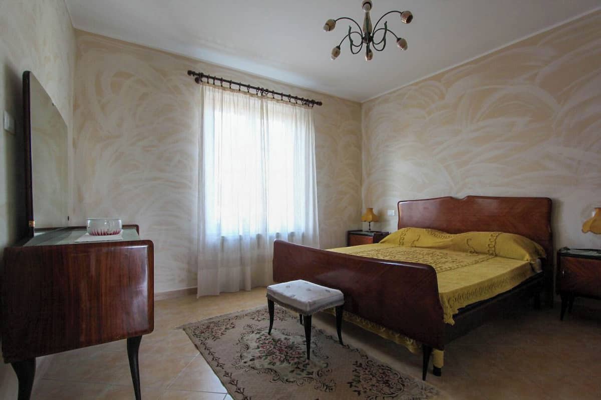 Ein Schlafzimmer mit Bett, Nachttisch, Stuhl, einem Fenster mit Vorhängen und einer Chandelier.