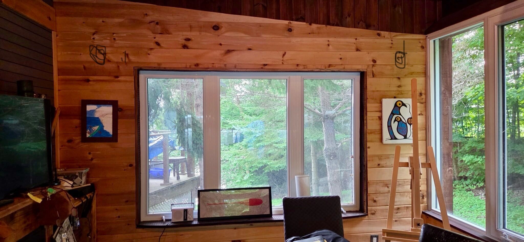 A cabin interior with a large window, wooden walls, a painting of birds, and a laptop on a desk.