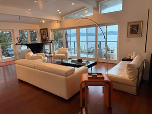 A spacious living room with a large window overlooking a body of water, a piano, and a modern coffee table. - Spacious waterfront 5-bedroom house in lovely Corte Madera with patio and pool (corte madera)