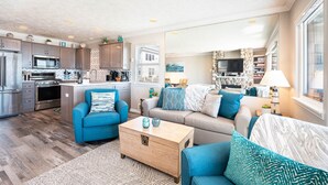Una sala moderna con un sofá azul, un sofá beige y una mesa de centro de madera. Hay una zona de cocina con microondas y refrigerador.