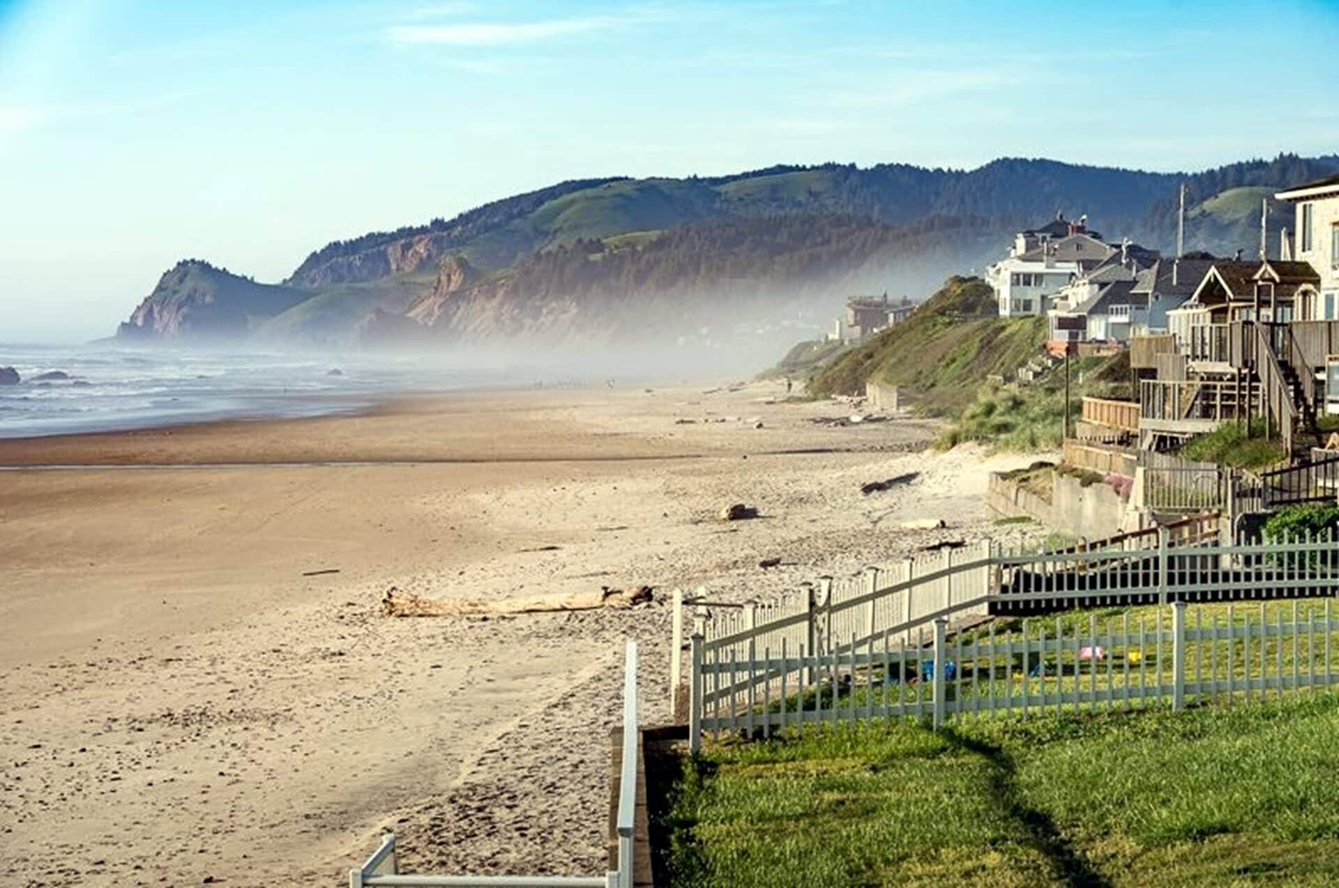 Una playa con una valla de madera, casas a la derecha y acantilados al fondo.