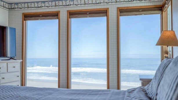 A bedroom with a large bed, a dresser, and a TV. It has three windows with a view of the ocean. - Logans Beachfront Lodge (Lincoln City)