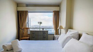 Quarto de hotel com uma cama grande, penteadeira, mesa de cabeceira e janela com cortinas.