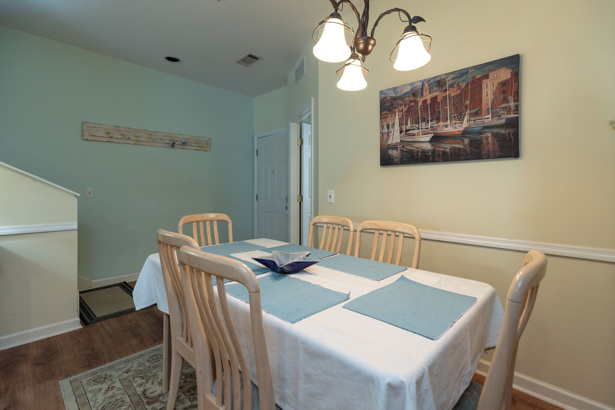 A dining room with a table set for four, wooden chairs, a white tablecloth, and a painting of a harbor scene on the wall.