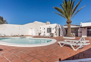 Area piscina con lettini, una palma e un edificio bianco con pavimento piastrellato.
