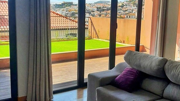 Un salon moderne avec un canapé gris, une table basse en verre et une vue sur le paysage urbain, visible à travers de grandes fenêtres.