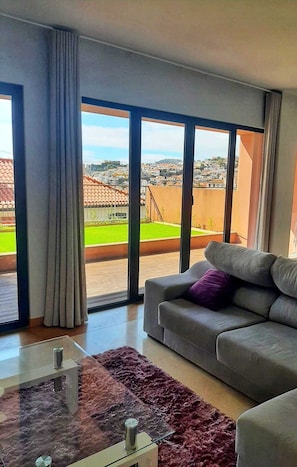 A modern living room with a grey sofa, a glass coffee table, and a view of a cityscape through large windows. - Swimming pool and private garden in the heart of Funchal (Funchal)