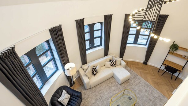 A modern living room with a sectional sofa, a black armchair, and a glass coffee table. There are large windows with curtains, a floor lamp, and a unique ceiling light fixture. - Luxury Church Conversion in Hale - Sleeps 4 (Altrincham)