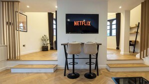 A modern living room with a flat-screen TV mounted on the wall displaying "NETFLIX". There are two bar stools with cushioned seats and black bases. The room has wooden flooring, a staircase leading to another level, and a potted plant on a side table. - Luxury Church Conversion in Hale - Sleeps 4 (Altrincham)