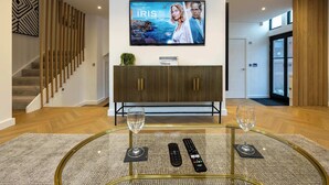 A modern living room with a glass coffee table, two wine glasses, and a flat-screen TV displaying 'The Iris Affair'. - Luxury Church Conversion in Hale - Sleeps 4 (Altrincham)