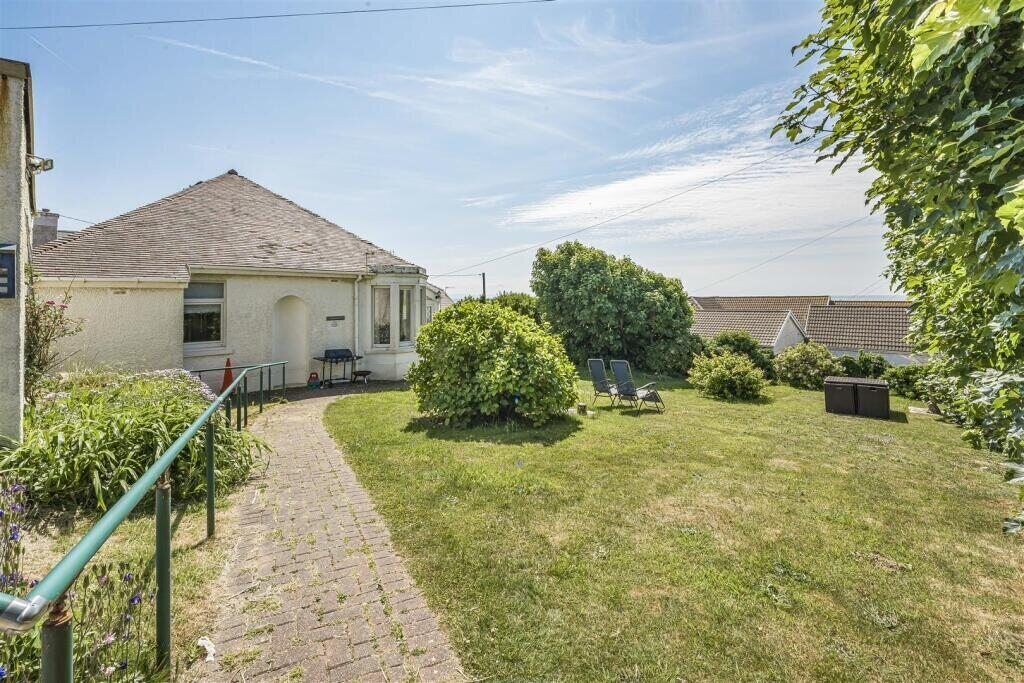 A residential property with a paved pathway leading to a house with a tiled roof. There is a well-maintained lawn with a large bush and a few outdoor chairs.