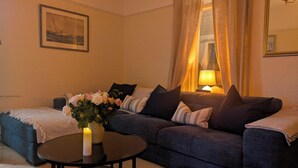 A cozy living room with a sofa, a small round table, a vase with flowers, a framed picture on the wall, and a lamp.