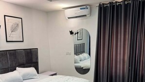 A modern bedroom with a large bed, a round mirror, an air conditioning unit, and a black ottoman. - Akira's  Homes (Abuja)