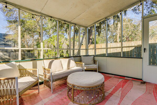 A covered outdoor seating area with wicker furniture and a round ottoman, large windows, and a view of trees and a fence. - Dogs Welcome! Savannah Escape w/ Fenced-in Yard (Savannah)