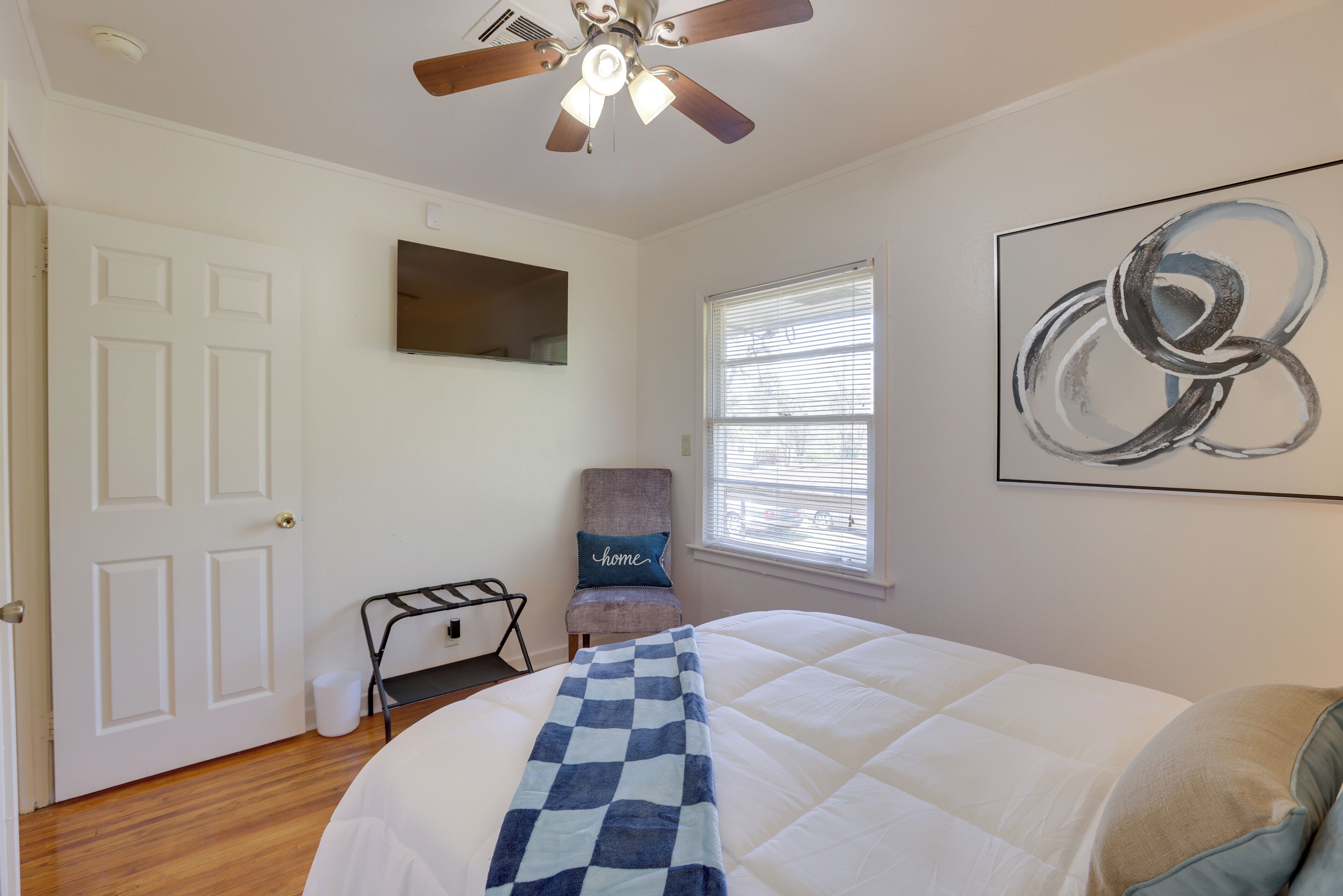A bedroom with a bed, ceiling fan, window, chair, and artwork on the wall.