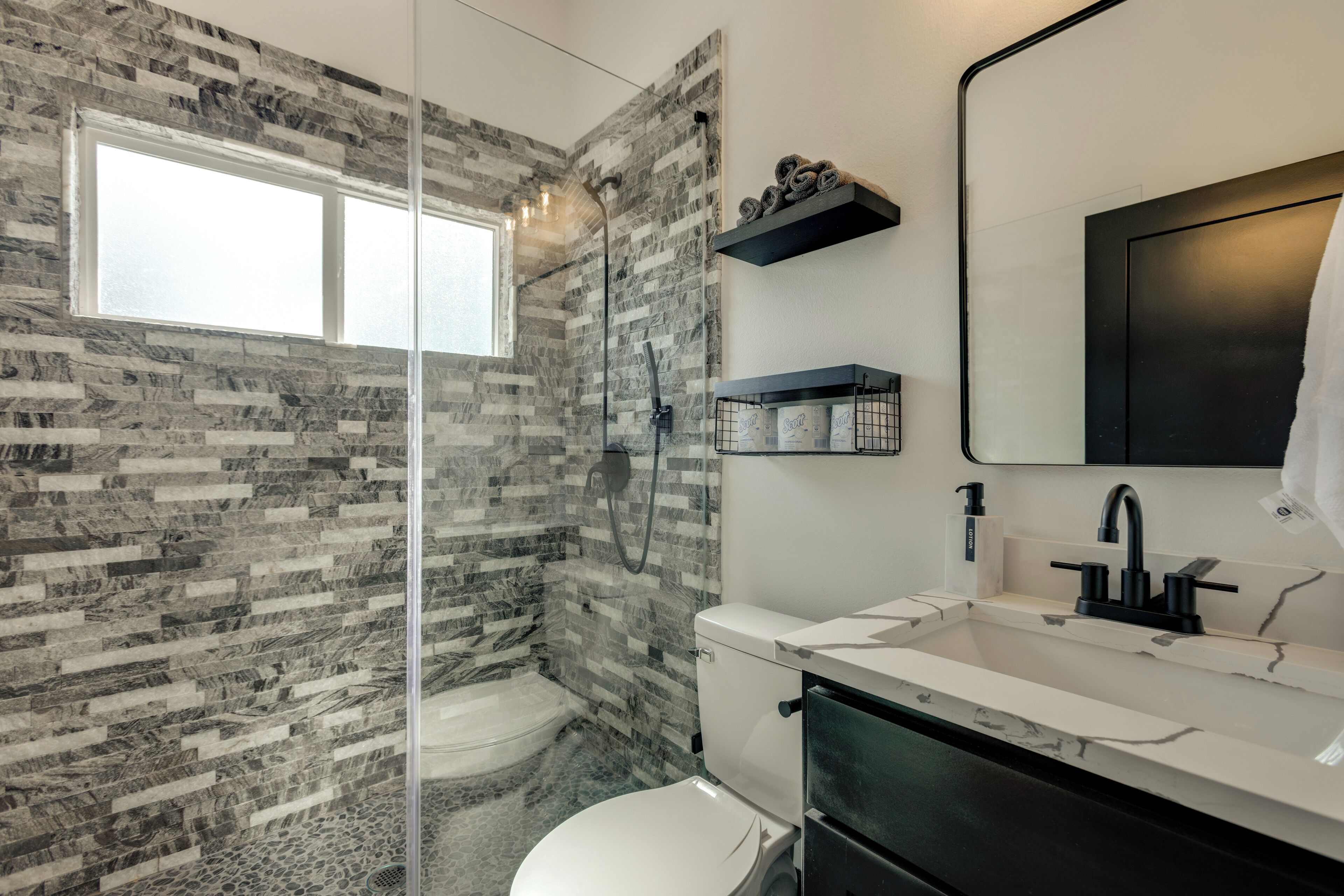 Un baño moderno con ducha envuelta en vidrio, inodoro blanco, lavabo blanco con grifo negro y estante negro sobre el lavabo.