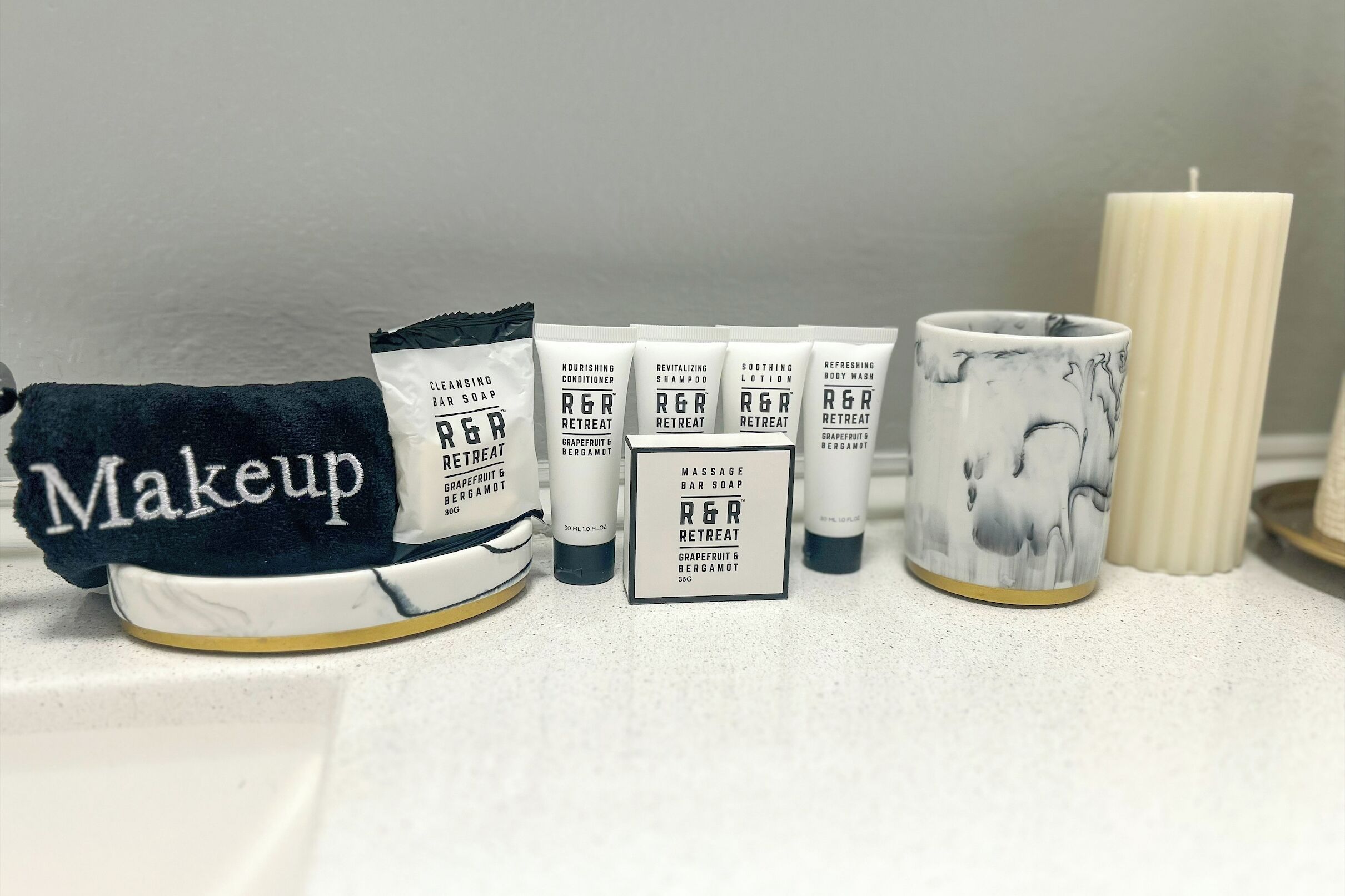 A bathroom counter with various R&R products including a towel, bar soap, shampoo, lotion, and a candle.
