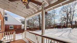 A covered porch with a wooden deck and white railings, overlooking a snow-covered yard with a brick house and wooden fences.